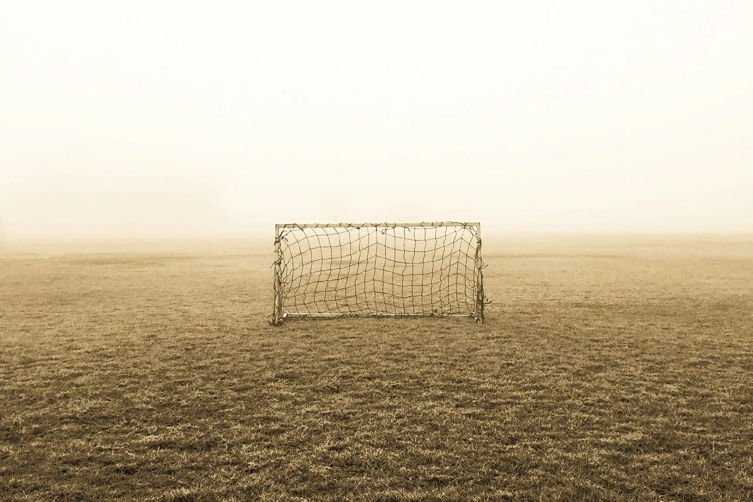 Kleines, leeres Fußballtor auf einem nebligen Spielfeld. Das Tor steht zentral auf einer weitläufigen, grasbewachsenen Fläche und wirkt durch den dichten Nebel im Hintergrund verlassen und ruhig.